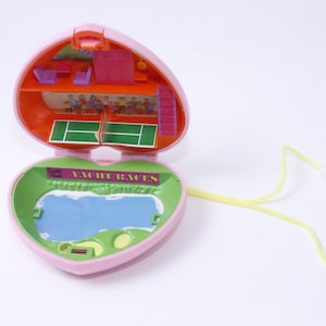 Könnte beinhalten: Ein rosa herzförmiges Spielzeug aus Plastik mit einer gelben Schnur. Das Spielzeug lässt sich öffnen und zeigt einen Miniatur-Tennisplatz und einen Pool mit dem Aufdruck "YACHT RACES" auf der Seite.