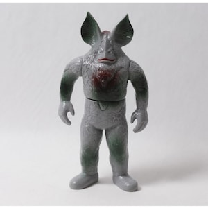May include: A gray humanoid monster figure with large, pointed ears, a textured body, and a red-painted chest and mouth area. The creature has a distinct, scaly skin pattern across its torso and legs, with dark green shading on the arms and sides.