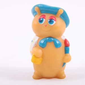 Op de afbeelding: Een vintage, bruin plastic speelgoedfiguur van een stripfiguur. De figuur heeft een blauwe hoed, een strik en accenten. Het houdt een gele tas en een penseel vast. De figuur heeft een lachend gezicht met blauwe ogen en rode accenten.