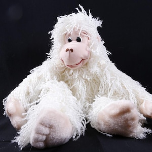 May include: A plush white yeti toy with a smiling face, pink nose, and pink hands and feet. The yeti has long, shaggy fur and is sitting with its legs spread out. The background is black.