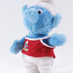 May include: A plush Smurf doll, featuring blue skin, a white hat, and white pants. The Smurf wears a red vest with a graphic. The doll has a smiling expression and is standing upright.