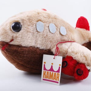 May include: A plush toy airplane in shades of tan, brown, and red. The toy has large black eyes, a red mouth, and white circular accents. A tag with the word "KAMAR" is attached.