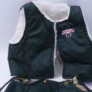 May include: Green quilted vest with a white sherpa lining and a "Teddy Ruxpin" logo patch. The vest has a zipper closure and two pockets. The vest is part of a set that includes matching pants.