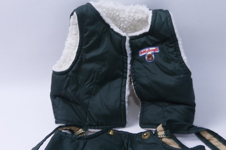 May include: Green quilted vest with a white sherpa lining and a "Teddy Ruxpin" logo patch. The vest has a zipper closure and two pockets. The vest is part of a set that includes matching pants.
