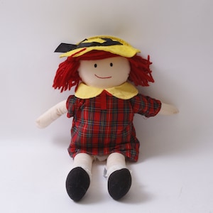 May include: A red-haired rag doll wearing a yellow and black hat, a red and black plaid dress, and black shoes.