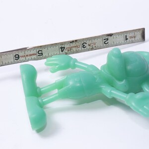 K&ouml;nnte beinhalten: Eine gr&uuml;ne Plastikfigur von Mickey Mouse, etwa 12,7 cm gro&szlig;, liegt auf einer wei&szlig;en Oberfl&auml;che. Ein Lineal befindet sich im Hintergrund zur Gr&ouml;&szlig;enbestimmung.