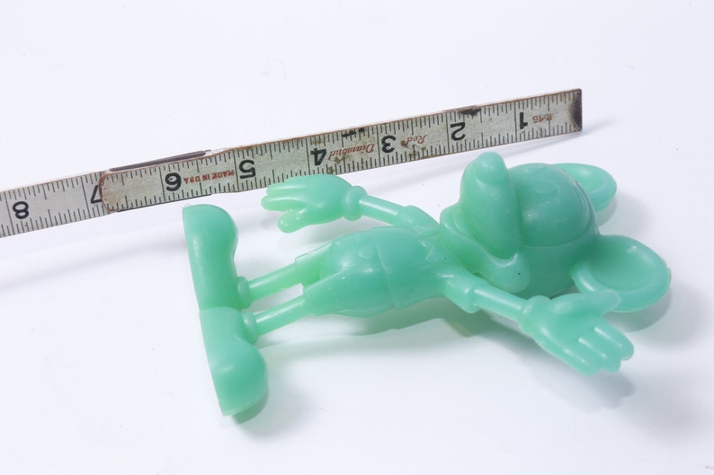 K&ouml;nnte beinhalten: Eine gr&uuml;ne Plastikfigur von Mickey Mouse, etwa 12,7 cm gro&szlig;, liegt auf einer wei&szlig;en Oberfl&auml;che. Ein Lineal befindet sich im Hintergrund zur Gr&ouml;&szlig;enbestimmung.