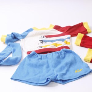 May include: A vintage children's athletic outfit in primary colors. The set includes a blue headband, blue shorts, a long-sleeved shirt with bicycle graphics, and red and yellow arm warmers. The shirt has red, blue, and yellow stripes.