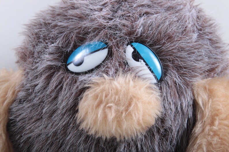 May include: A brown and gray furry toy with blue eyes. The toy has a brown fuzzy nose and a white mouth.