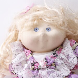 May include: A vintage Cabbage Patch Kid doll with blonde curly hair, blue eyes, and a pink bow. The doll is wearing a white lace collar and a floral dress with pink, purple, and yellow flowers.