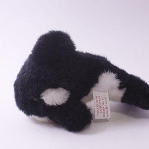 May include: A black and white orca whale plush toy. The soft toy has a furry texture and white markings. A tag is attached to the toy.