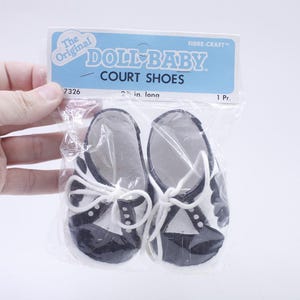 May include: A pair of Doll-Baby court shoes in a clear plastic package. The shoes are black and white with white laces. The package has a blue and white label that reads "DOLL-BABY COURT SHOES". The shoes are 2.5 inches long.