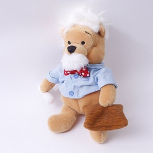 May include: A plush Winnie the Pooh bear toy wearing a blue shirt with red polka dots and a red bow tie. The bear has a white beard and white hair. The bear is holding a brown wooden object.