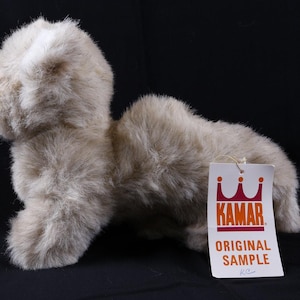 May include: A plush, light brown stuffed animal in a quadrupedal pose. The toy has a shaggy texture and a tag that reads "KAMAR ORIGINAL SAMPLE". The background is black.