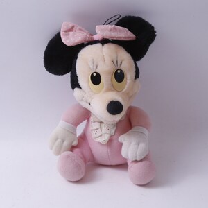 May include: A soft toy Minnie Mouse doll, seated and wearing a pink outfit with a matching bow. The doll has black ears, a cream-coloured face, and large yellow eyes. The bow features white polka dots.