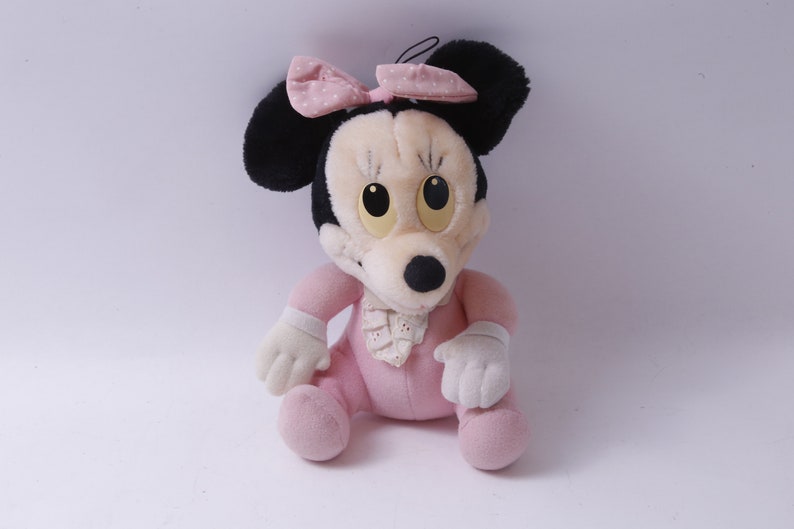 May include: A soft toy Minnie Mouse doll, seated and wearing a pink outfit with a matching bow. The doll has black ears, a cream-coloured face, and large yellow eyes. The bow features white polka dots.