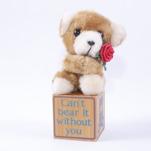 Op de afbeelding: Een bruine teddybeer met een rode roos in zijn poot zit op een houten blok met de tekst "Can't bear it without you".