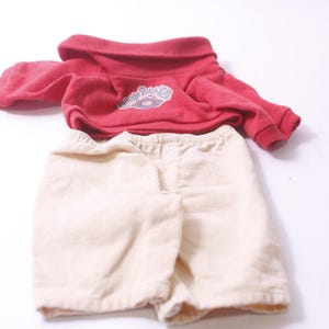 May include: A red hooded sweatshirt and cream-colored corduroy shorts. The sweatshirt has a small logo on the front. The shorts have an elastic waistband. This outfit is suitable for a doll.