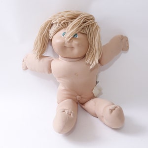 Può includere: Una bambola vintage Cabbage Patch Kid con capelli di lana biondi e occhi azzurri. La bambola è seduta con le gambe incrociate e le braccia distese.