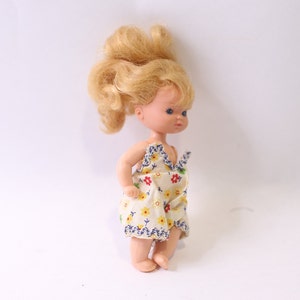 May include: A small plastic doll with blonde curly hair wearing a white dress with a floral pattern.