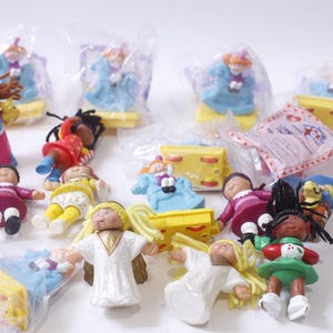 May include: A collection of colorful plastic dolls, some wearing dresses and others wearing pants, with various accessories. The dolls are packaged in clear plastic bags and are arranged on a white surface.