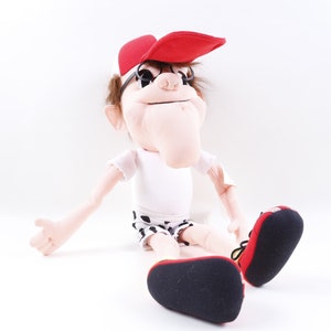 May include: A plush puppet with a red cap, black glasses, and white shirt. The puppet is wearing black and white polka dot shorts and red and black shoes. The puppet is sitting on a white surface.