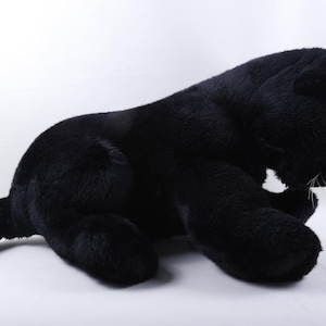 May include: A plush toy shaped like a black panther with soft, dark fur, rounded ears, and a long tail. The stuffed animal features small, dark eyes, white whiskers, and a small, light-coloured nose, depicted in a resting, lying-down position.