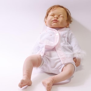 May include: A lifelike baby doll, dressed in a white outfit and a pink-edged jacket. The doll has closed eyes and brown hair. The jacket has a textured pattern and button closure. The doll is lying down.