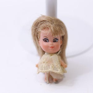May include: A small vintage doll with blonde hair and large brown eyes. The doll is wearing a light yellow dress with a floral pattern. The doll is standing on a white surface.