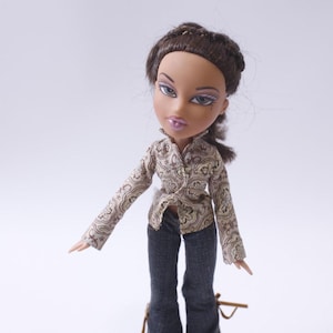 Puede incluir: Una muñeca de pelo castaño con ojos marrones y una camisa de estampado marrón claro y negro. La muñeca lleva pantalones vaqueros azul oscuro y tiene una trenza marrón claro en el pelo.