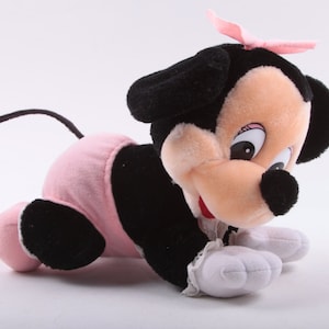 May include: A plush toy of a black and pink Minnie Mouse crawling on a white background. The toy has a pink bow on its head and is wearing a pink diaper.