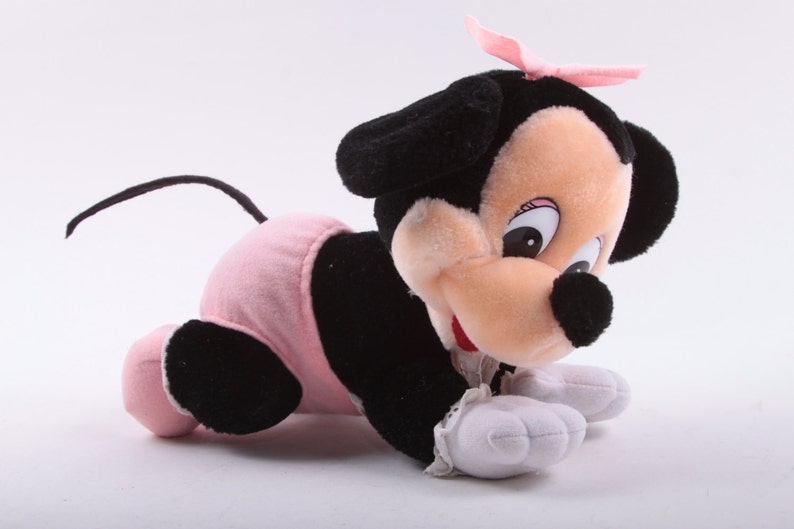 May include: A plush toy of a black and pink Minnie Mouse crawling on a white background. The toy has a pink bow on its head and is wearing a pink diaper.