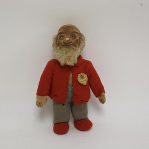 May include: A vintage doll with a painted face, a beige beard, and a red jacket. The doll is wearing gray pants and red shoes. A small, round tag is pinned to the jacket. The doll is standing against a white background.