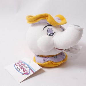 Può includere: Un peluche a forma di personaggio teiera con viso bianco, palpebre viola e un bordo giallo arricciato. Il giocattolo presenta un beccuccio prominente, una consistenza morbida e un'etichetta di carta bianca con il testo The Disney Store.