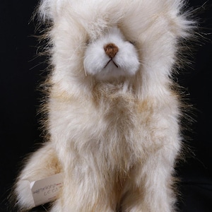 May include: A plush toy animal with long, shaggy, cream and light brown faux fur. The toy has a white face, a small brown nose, and a visible white tag on its side with the text Birmingham printed in black ink against a solid black background.