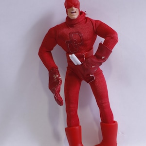 May include: A red Daredevil action figure with a white mask and a red suit. The figure is holding two white sticks in its hands. The figure is standing on a white background.