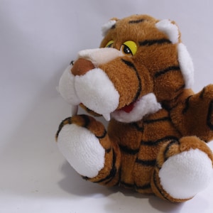 Könnte beinhalten: Ein braunes und weißes Plüschtiger-Spielzeug mit schwarzen Streifen. Der Tiger hat ein weißes Gesicht mit einer schwarzen Nase und gelben Augen. Das Spielzeug sitzt auf einem weißen Hintergrund.