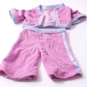May include: A two-piece pink velour outfit for a doll or small child. The set includes a short-sleeved jacket and matching cropped pants, both trimmed with light purple fabric. The jacket has a front closure.