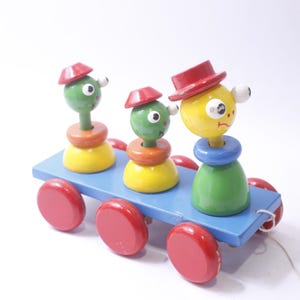 May include: A wooden pull toy featuring three colourful characters on a blue platform with red wheels. The characters have green and yellow bodies, red hats, and googly eyes.