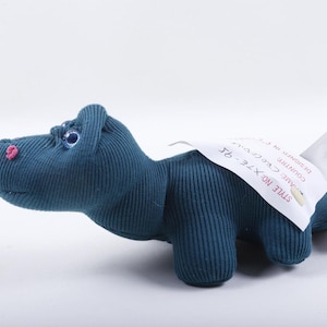 May include: A teal ribbed fabric dinosaur-shaped tissue dispenser with a small pink nose and blue eyes. A white tag is attached to the back, featuring the text STYLE NO: X7E-95, along with other partially visible printed information.