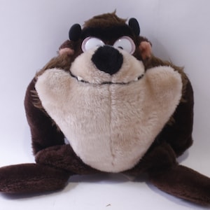 Può includere: Un giocattolo di peluche marrone e beige del personaggio dei cartoni animati Taz, il diavolo della Tasmania. Il giocattolo è seduto con le gambe distese e ha un ampio sorriso.