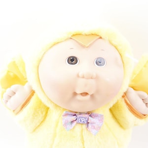 May include: A vintage Cabbage Patch Kids doll wearing a yellow hooded outfit with a light purple bow. The doll has one brown eye and one blue eye. The doll's outfit has a soft, plush texture.