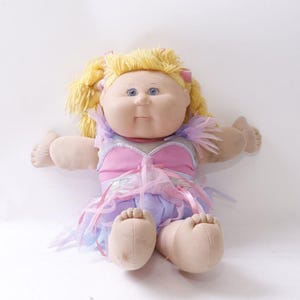 May include: A vintage Cabbage Patch Kids doll with blonde hair and blue eyes. The doll is wearing a pink and purple dress with a tulle skirt and has its arms outstretched. The doll's skin is a light beige color.