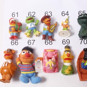 May include: A collection of Sesame Street character figurines, including Bert, Ernie, Oscar the Grouch, and others. The figurines are colorful and detailed, with some wearing costumes. The image is taken against a white background with numbers.