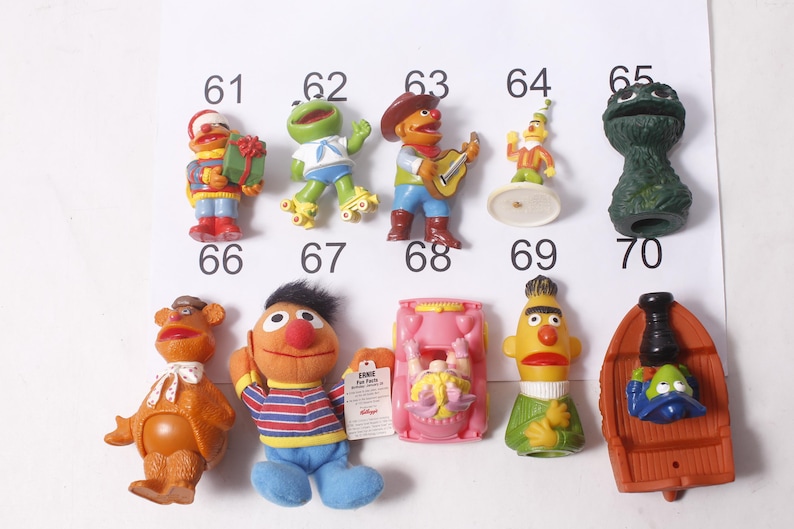 May include: A collection of Sesame Street character figurines, including Bert, Ernie, Oscar the Grouch, and others. The figurines are colorful and detailed, with some wearing costumes. The image is taken against a white background with numbers.