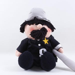 May include: A plush doll depicting a police officer, featuring a black uniform with a gold star badge, a gray hat, and a black mustache. The doll has a light skin tone and is holding a gray baton.
