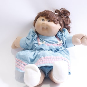 Puede incluir: Una muñeca Cabbage Patch Kid con cabello castaño rizado que lleva un vestido azul y rosa. La muñeca tiene un vestido azul claro con ribetes rosas y calcetines blancos.