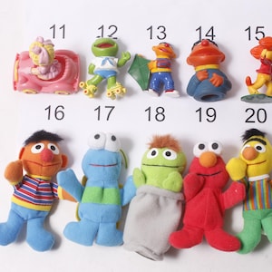 May include: A collection of Sesame Street character figurines and plush toys. The image includes figures of Elmo, Bert, Ernie, Cookie Monster, and others. The toys are colorful and feature recognizable character designs.