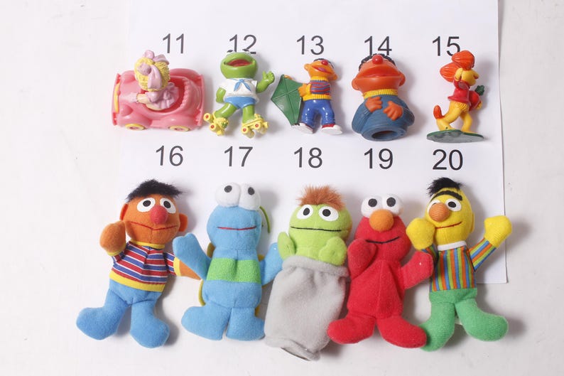 May include: A collection of Sesame Street character figurines and plush toys. The image includes figures of Elmo, Bert, Ernie, Cookie Monster, and others. The toys are colorful and feature recognizable character designs.