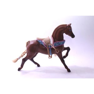 以下が含まれることがあります： 三つ編みの尾と鞍が付いた茶色の玩具の馬。鞍はタン色で、青とピンクの模様のトリムが付いています。馬は走っているかのようにポーズをとっています。